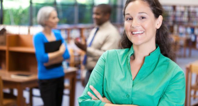 A woman in a library smiling at the camera and two adults talk behind her.